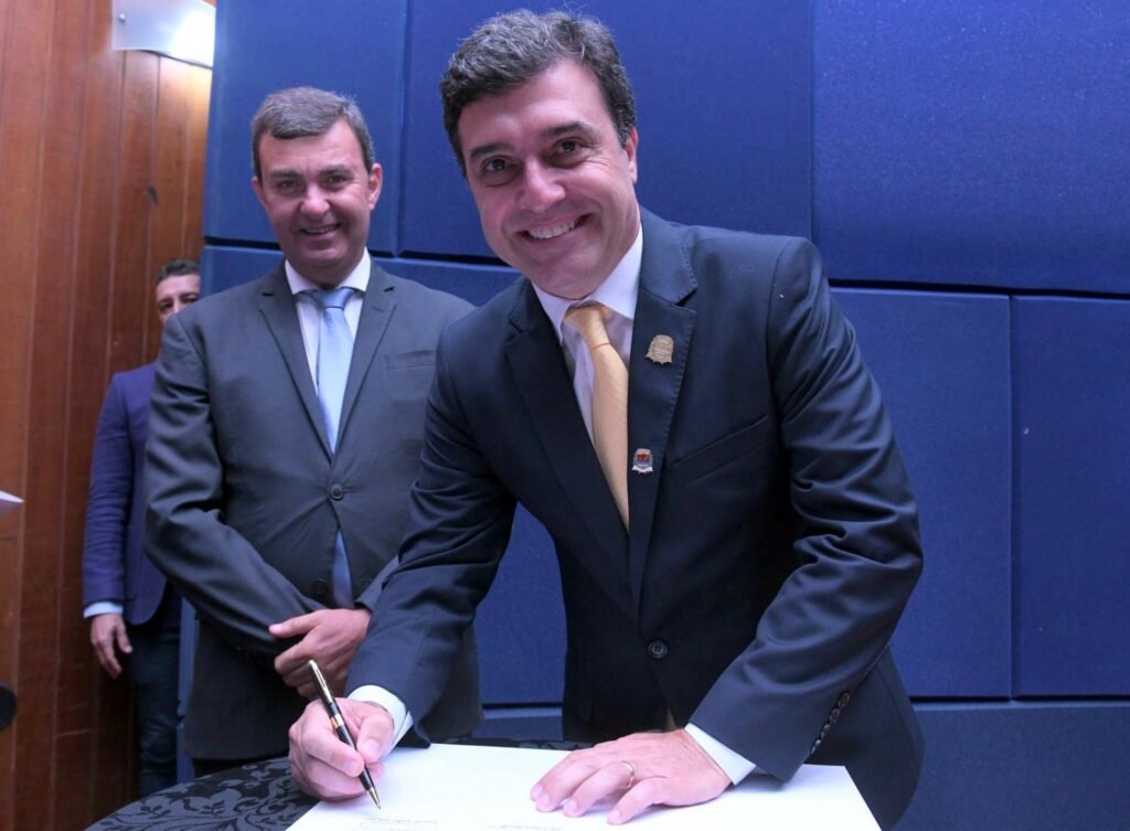 a imagem colorida mostra o prefeito Dr. Custódio Tavares Dias Neto assinando o Termo de Posse no Auditório da Prefeitura. Ele está em pé no palco, de frente para o público, sorrindo, assinando o documento que está sobre uma mesa redonda com toalha azul. Ao fundo, o ex-prefeito Nilson Alcides Gaspar, também sorrindo.