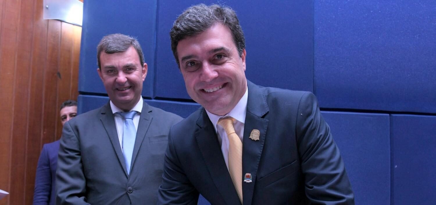 magem colorida mostra o prefeito Dr. Custódio Tavares Dias Neto assinando o Termo de Posse no Auditório da Prefeitura. Ele está em pé no palco, de frente para o público, sorrindo, assinando o documento que está sobre uma mesa redonda com toalha azul. Ao fundo, o ex-prefeito Nilson Alcides Gaspar, também sorrindo.