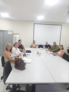 Uma mesa com os membros do conselho do Comdefi, Daniela do Funssol que organizou a reunião e a Débora Ciriaco, intérprete de libras e Coordenadora de Acessibilidade da Secretaria de Comunicação. Fim da descrição.