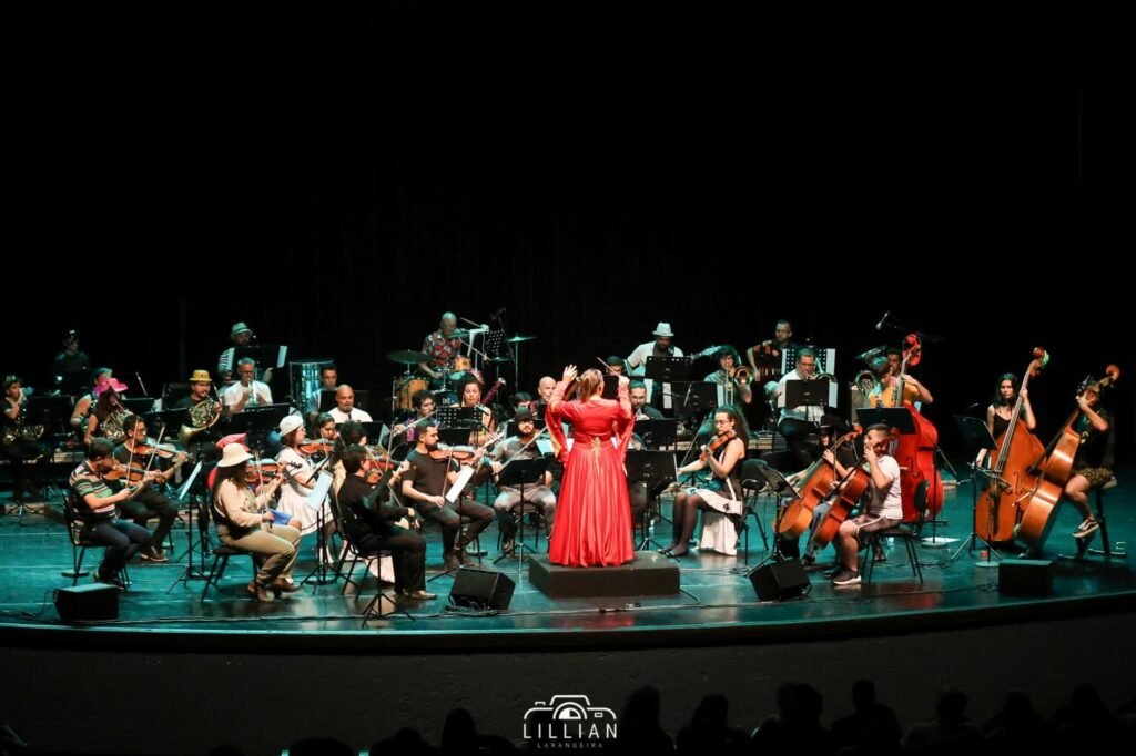 Na foto a maestrina regendo a orquestra. Em cima do palco músicos com instrumentos e partituras e a frente a maestrina com uma batuta regendo..