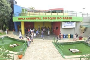 Foto colorida com a fachada da Escola Ambiental Bosque do Saber. Muitas pessoas no hall de entrada, famílias, crianças e adultos.