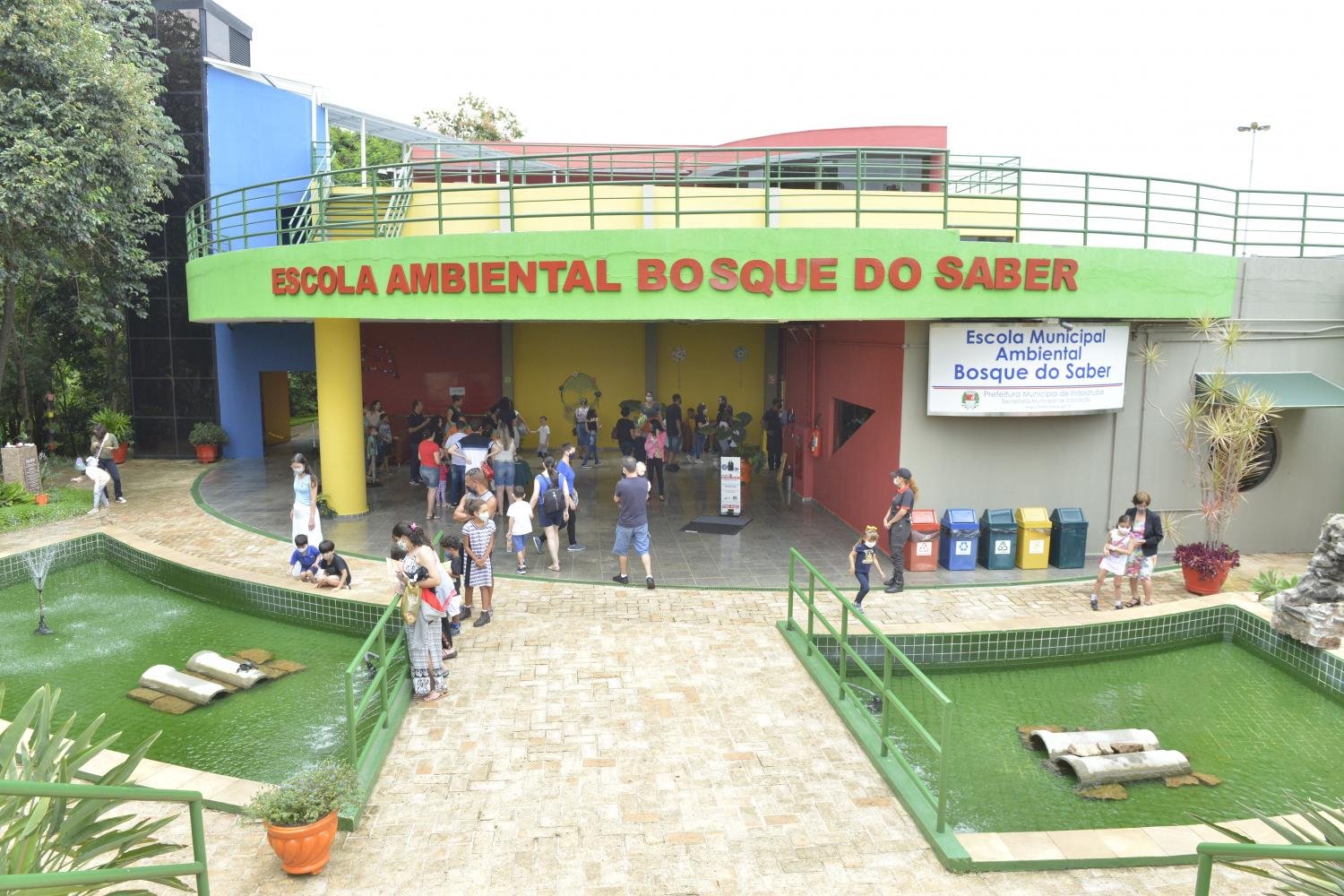 Foto colorida com a fachada da Escola Ambiental Bosque do Saber. Muitas pessoas no hall de entrada, famílias, crianças e adultos.