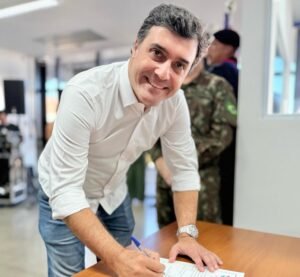 Foto colorida mostra o prefeito Dr. Custódio Tavares assinando o Termo de Posse como presidente da Junta Militar de Indaiatuba. Ele está em pé, levemente curvado sobre o documento na mesa, segurando uma caneta azul e sorrindo, olhando para a câmera. Ele veste uma camisa branca de mangas longas e uma calça jeans. Ao fundo, um homem com uniforme camuflado do Exército.