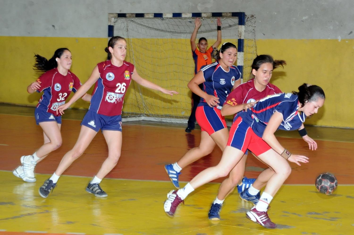 Seis atletas de handebol em quadra jogando e disputando a bola.