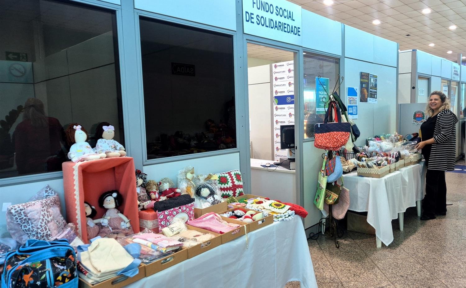 Foto colorida com professora Edileny Gomes de França, e produtos que estão a venda em frente ao Funssol, dentro da Prefeitura, expostos em cima de mesas, bonecas, panos de prato, bolsas e muito mais.