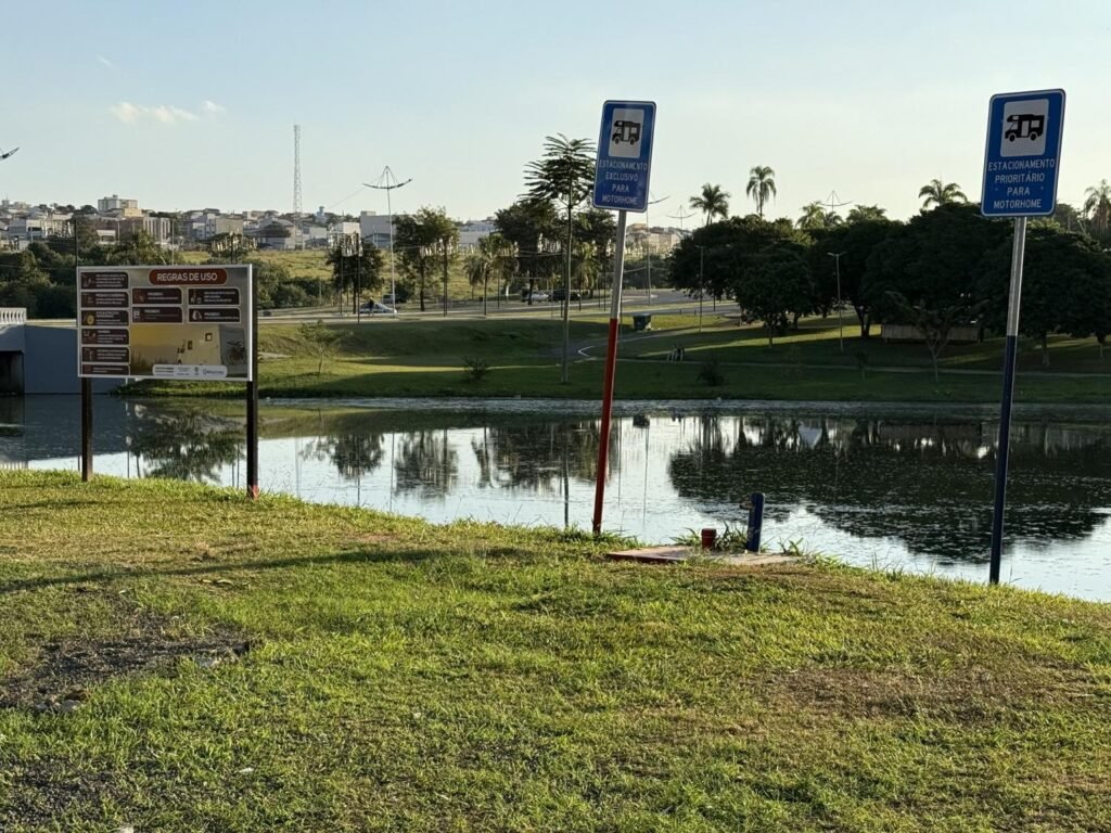 A foto colorida mostra o espaço reservado para estacionamento de motorhome no Parque Ecológico, próximo à pista de bicicross, com duas placas azuis indicando o ponto de estacionamento, ao lado de uma placa colorida maior, com as regras do local. As placas estão na margem do lago e ao fundo é possível ver árvores e, do lado esquerdo, alguns imóveis do Jardim Regina.