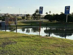 A foto colorida mostra o espaço reservado para estacionamento de motorhome no Parque Ecológico, próximo à pista de bicicross, com duas placas azuis indicando o ponto de estacionamento, ao lado de uma placa colorida maior, com as regras do local. As placas estão na margem do lago e ao fundo é possível ver árvores e, do lado esquerdo, alguns imóveis do Jardim Regina.
