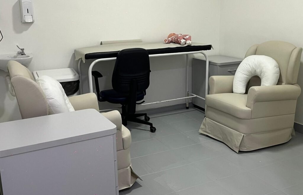 A imagem mostra uma maca para atendimento ao centro e no fundo da sala, e duas poltronas com almofada de amamentação em cada lado.