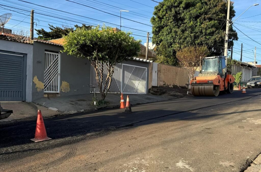 A foto colorida mostra um trecho da rua Vicente de Genaro com parte do asfalto já recapeado e separado por cones laranja e branco. Do lado direito, uma motoniveladora na cor laranja trafega na parte do asfalto recapeado. Ao fundo, os muros de residências da rua e árvores plantadas na calçada.