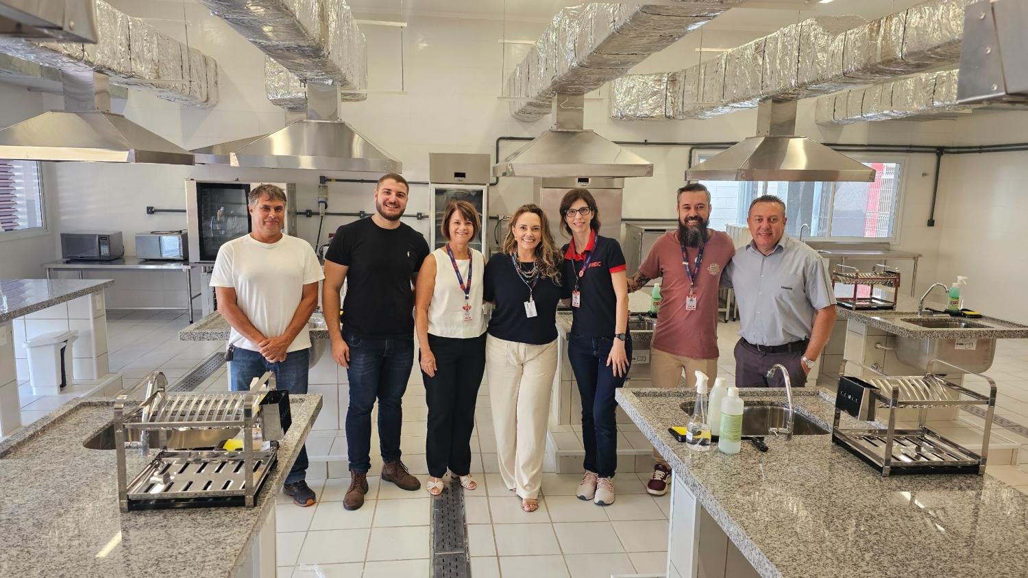 te pessoas estão em pé, sorrindo, lado a lado, em uma cozinha didática moderna com bancadas de granito e coifas de aço inox. Ao fundo, é possível ver fornos e micro-ondas. Da esquerda para a direita estão Rogério Brolo e André Antal, representantes do Ponto Verde. A superintendente Rita de Cassia Trasferetti, Mariela Silva Ferraz - Coordenadora dos Cursos de Gestão, Janaina Madruga Morais Ferreira - Coordenadora de Nutrição e Gastronomia e Samuel Moreira da Silva - Vice-Diretor Pedagógico, representando a Fiec e Ivan Nogueira – Representante Semurb.