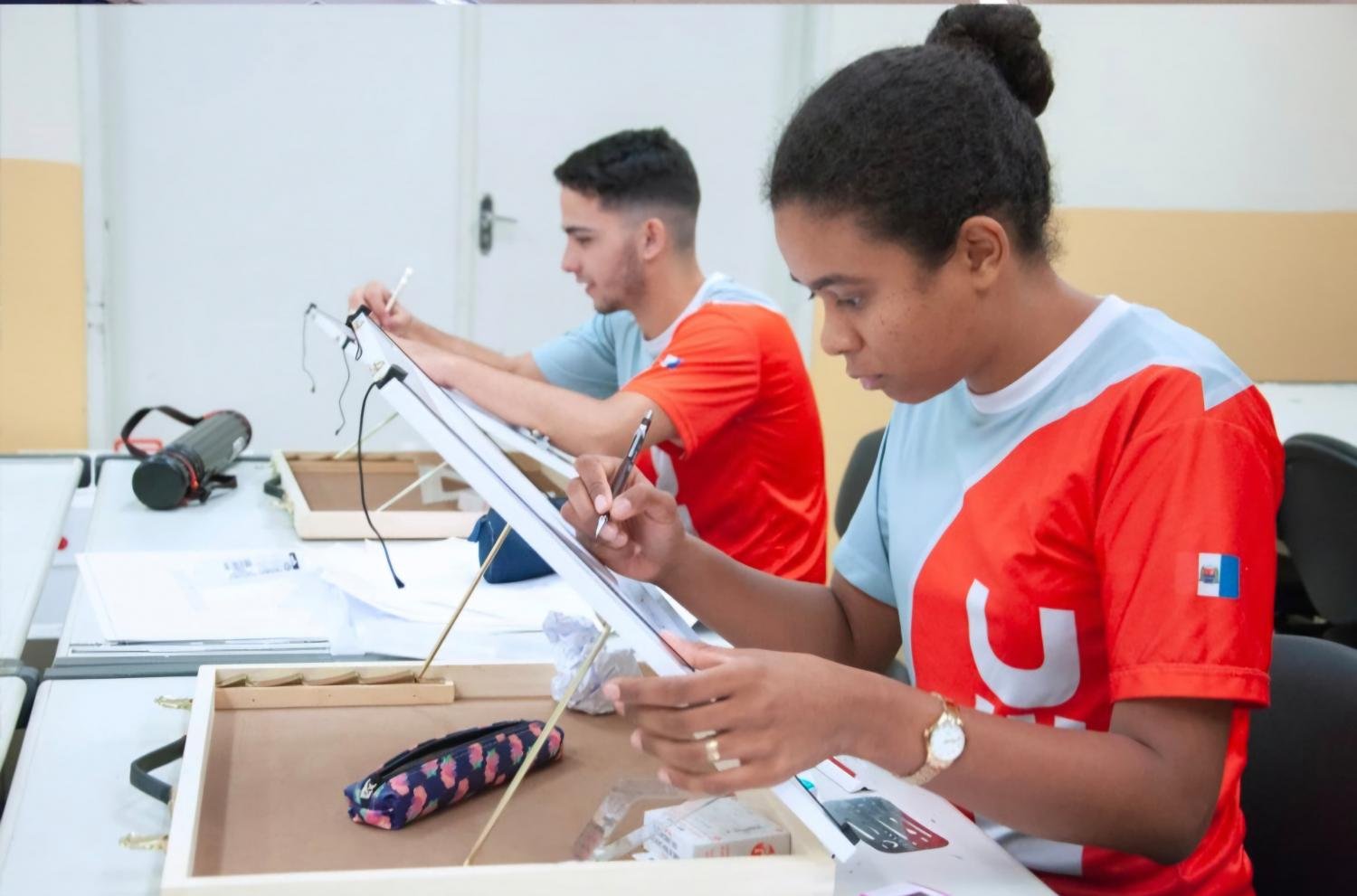 Dois jovens, uma menina e um menino, com uniforme vermelho da Fiec, olhando para tablets e estudando.
