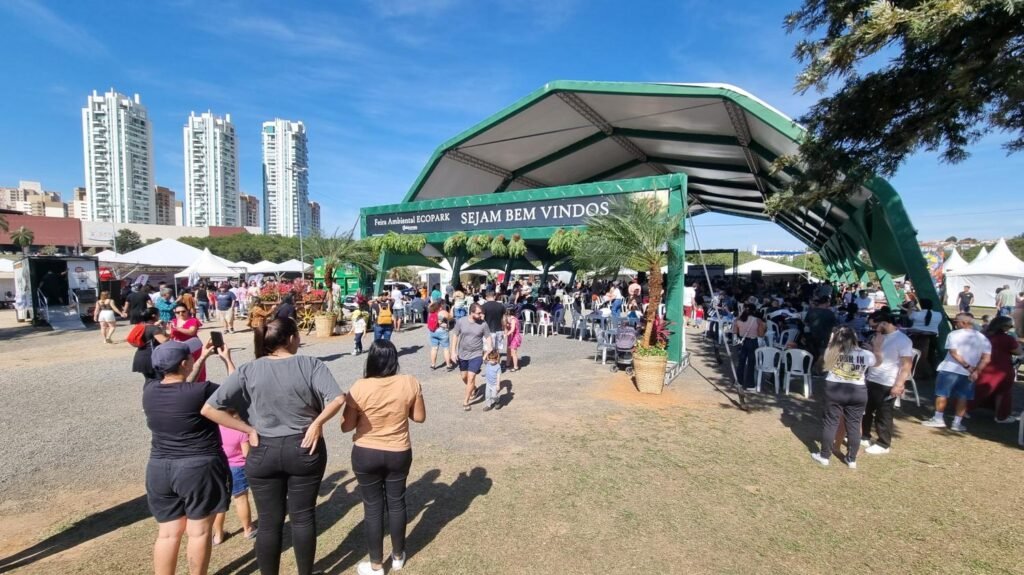 Foto colorida mostra a entrada principal da Feira Ambiental EcoPark, com a frase SEJAM BEM VINDOS no painel verde da entrada. Muitas pessoas estão caminhando, entrando no evento pela Tenda principal, com estrutura verde e cobertura branca. Ao fundo, três prédios se destacam com um céu azul de fundo.